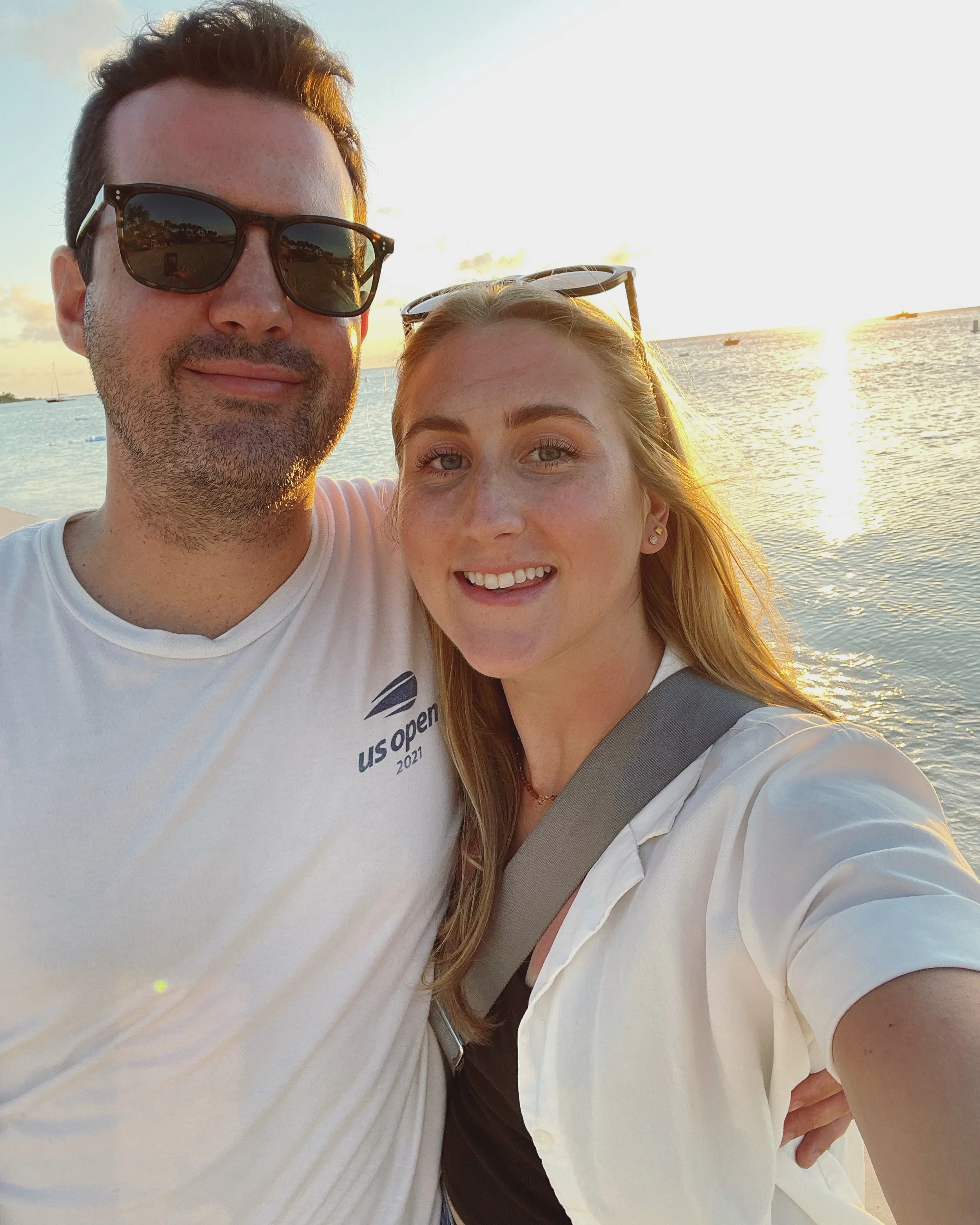 A smiling couple taking a selfie at sunset by the water, with boats in the distance.