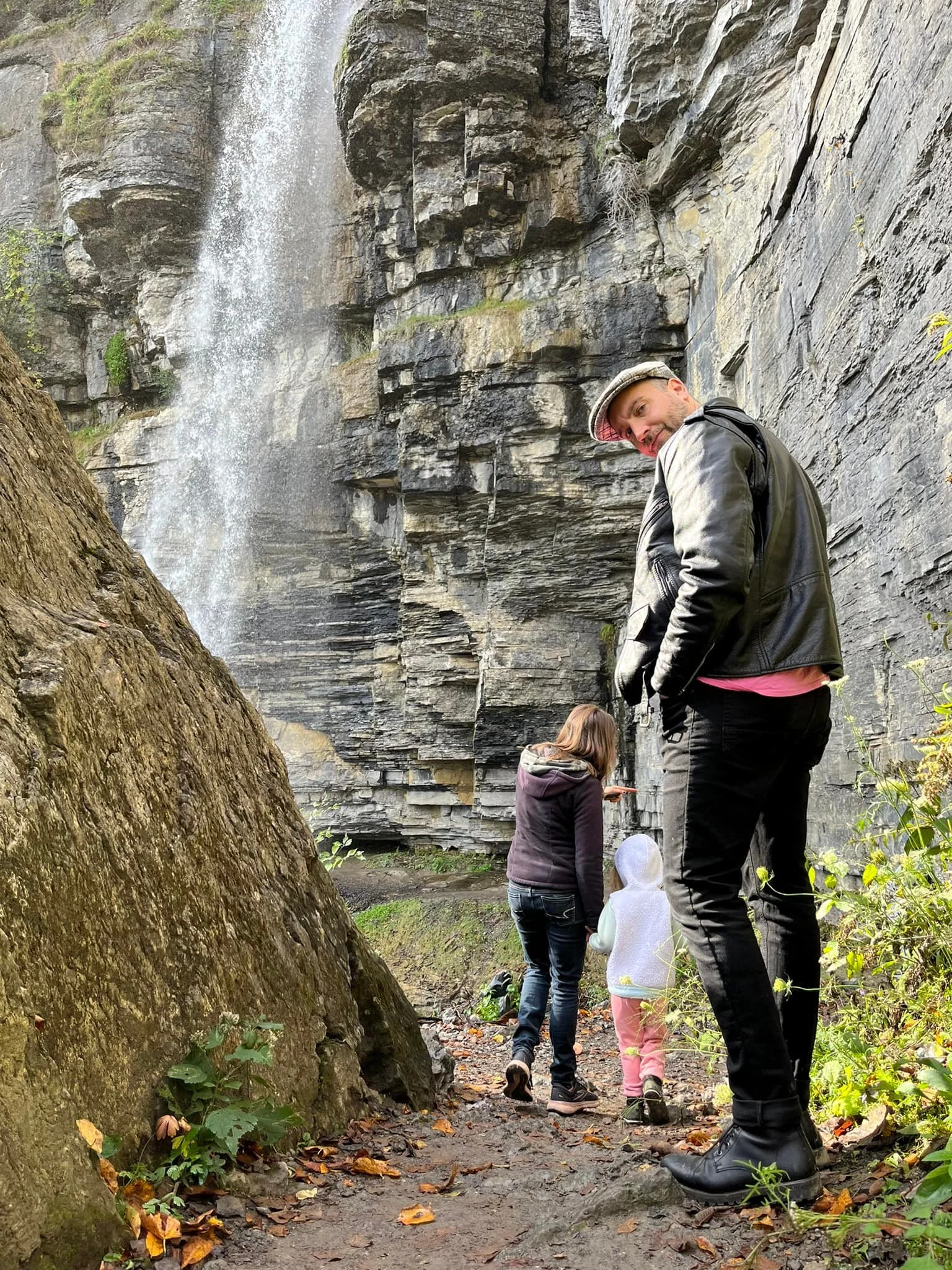 A man with a leather jacket and hat looking at the camera while standing on a trail in front of a waterfall. Two women and a child are walking ahead of him, exploring near the water.