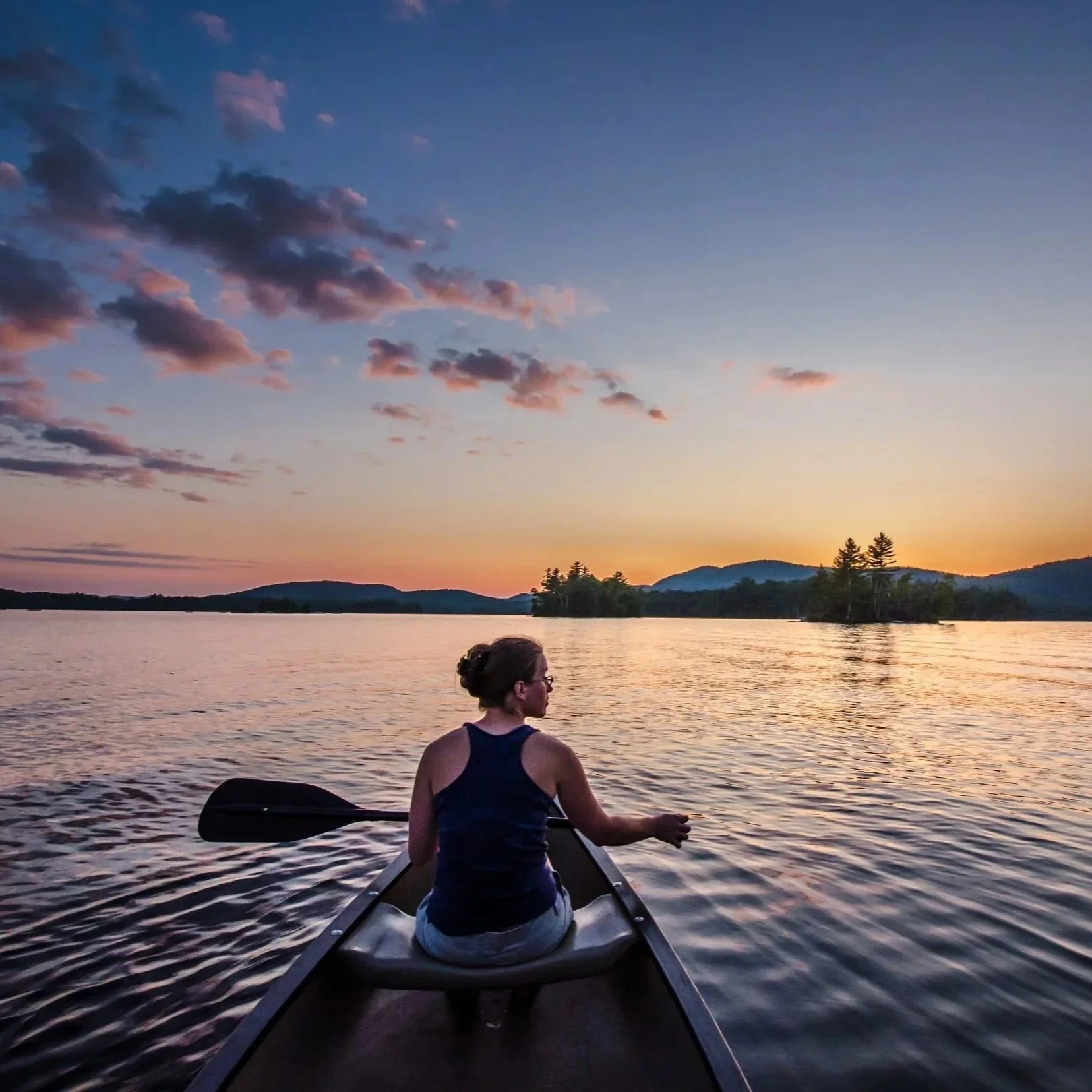 Shelly Cihan, Rooted Rock Marketing founder, paddles a canoe at sunset on an Adirondack lake, with mountains and forest in the background.