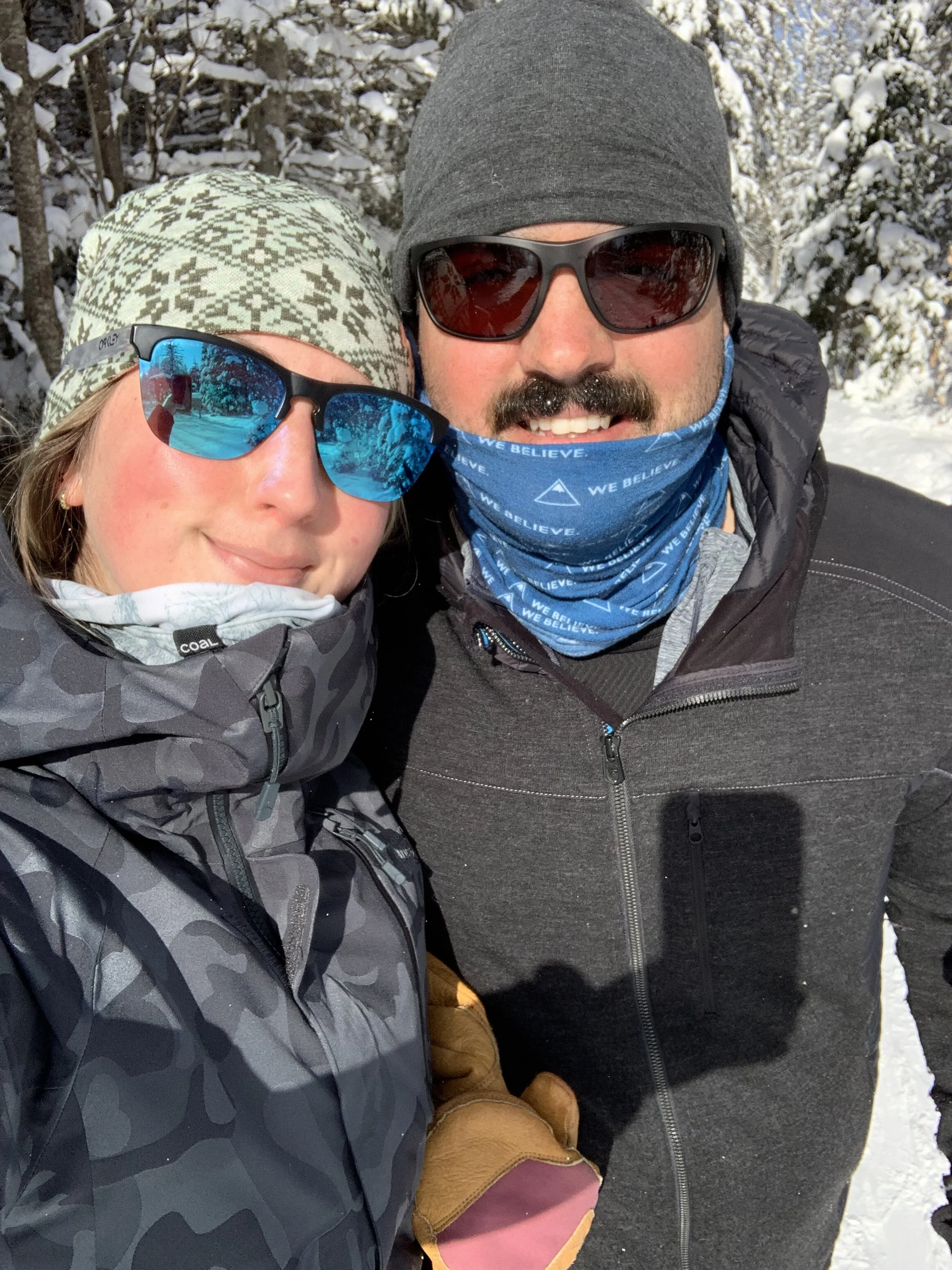 A woman and a man wearing winter outdoor gear, sunglasses, and beanies taking a selfie outdoors in a snowy landscape with trees covered in snow.