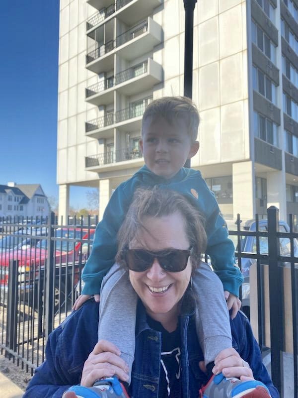 A woman with sunglasses carrying a young boy on her shoulders outdoors near a building and parking lot.