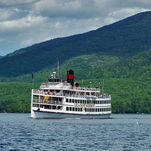 A large white steam-powered paddleboat with three levels floats on Lake George, with the Adirondacks in the background.