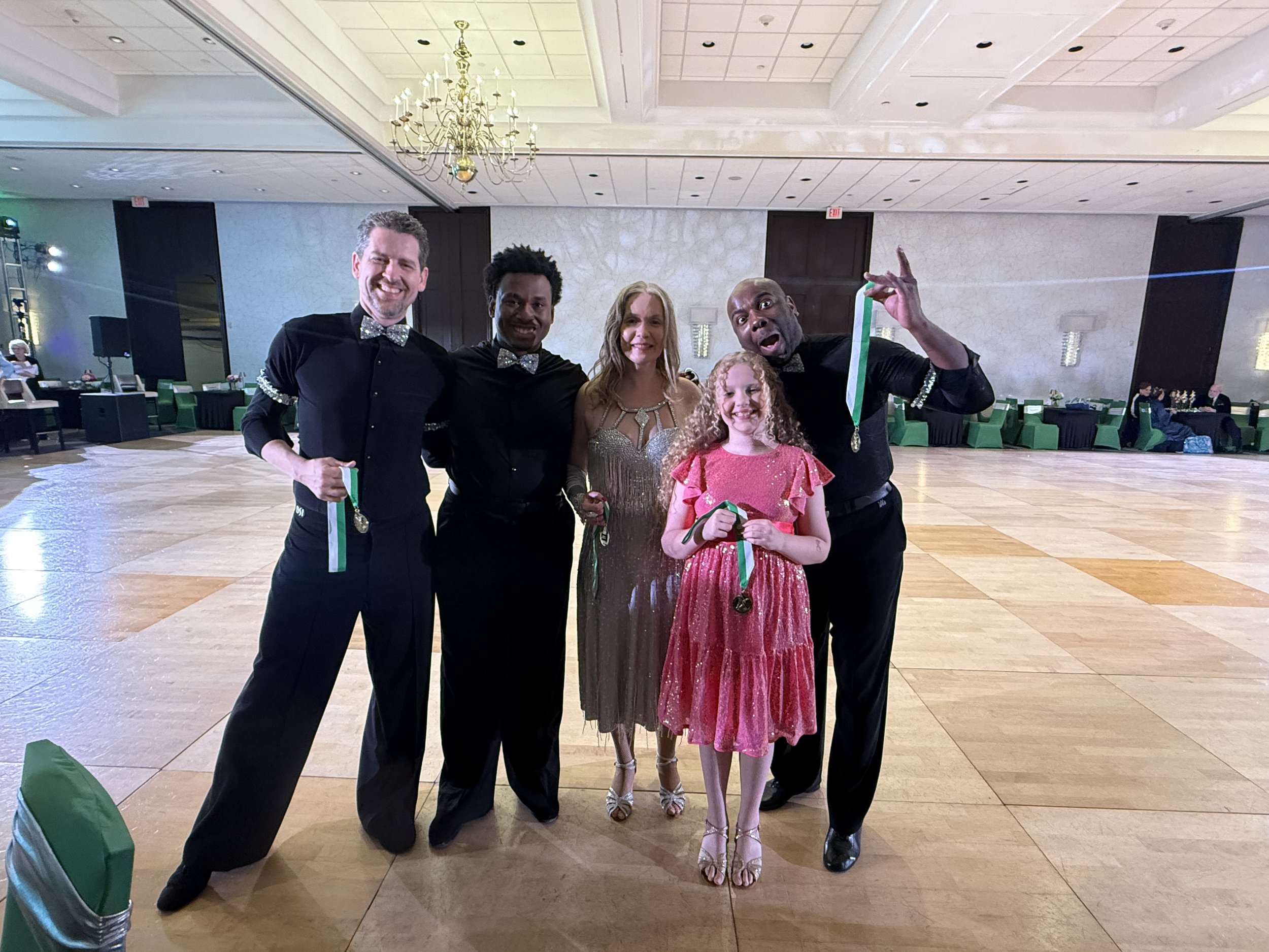 Group of five people posing in a ballroom holding medals, wearing formal attire.