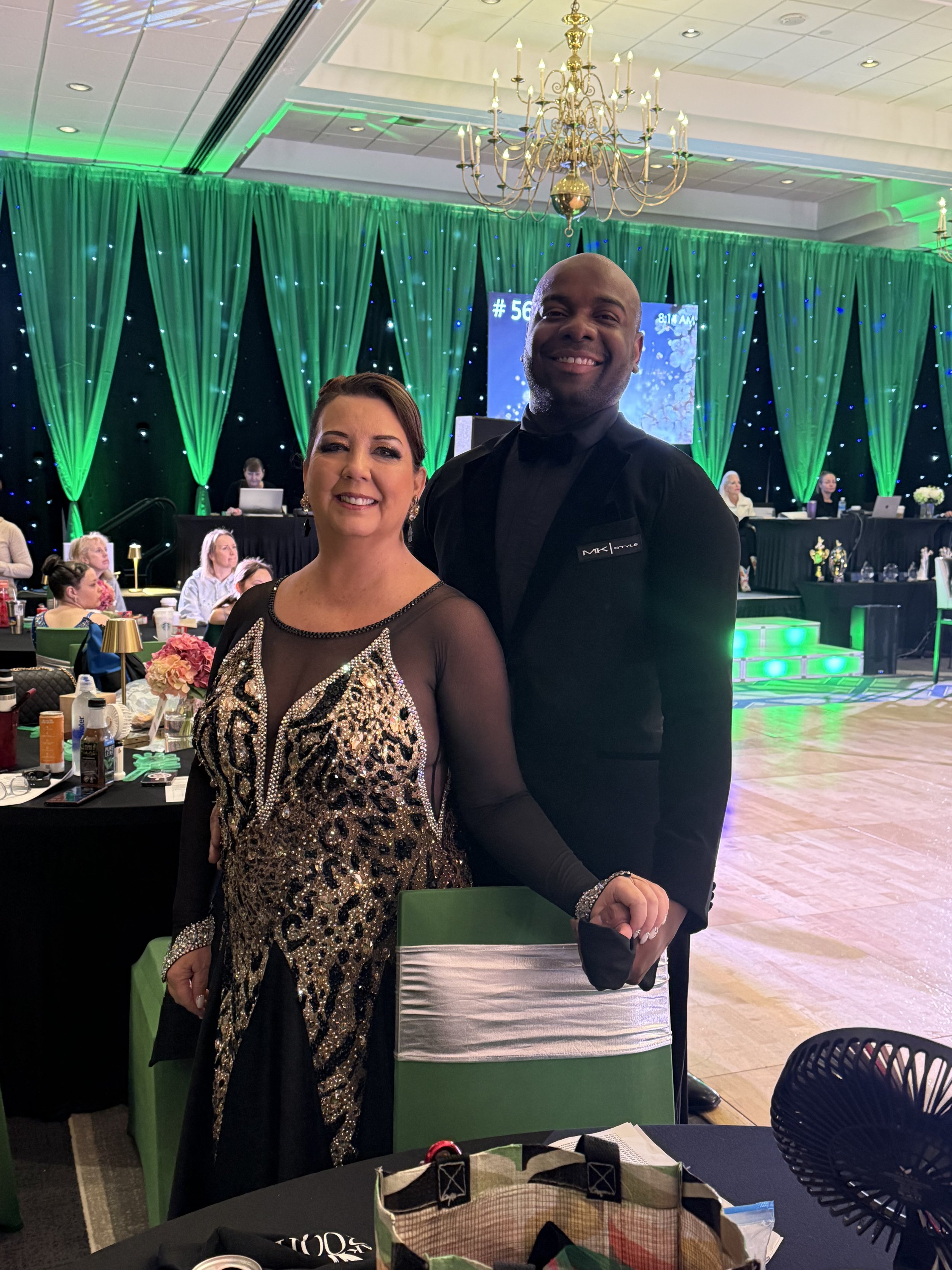 A man and woman in formal dance attire smiling, standing in a ballroom decorated with green curtains and chandeliers. Other people are seated at tables in the background.