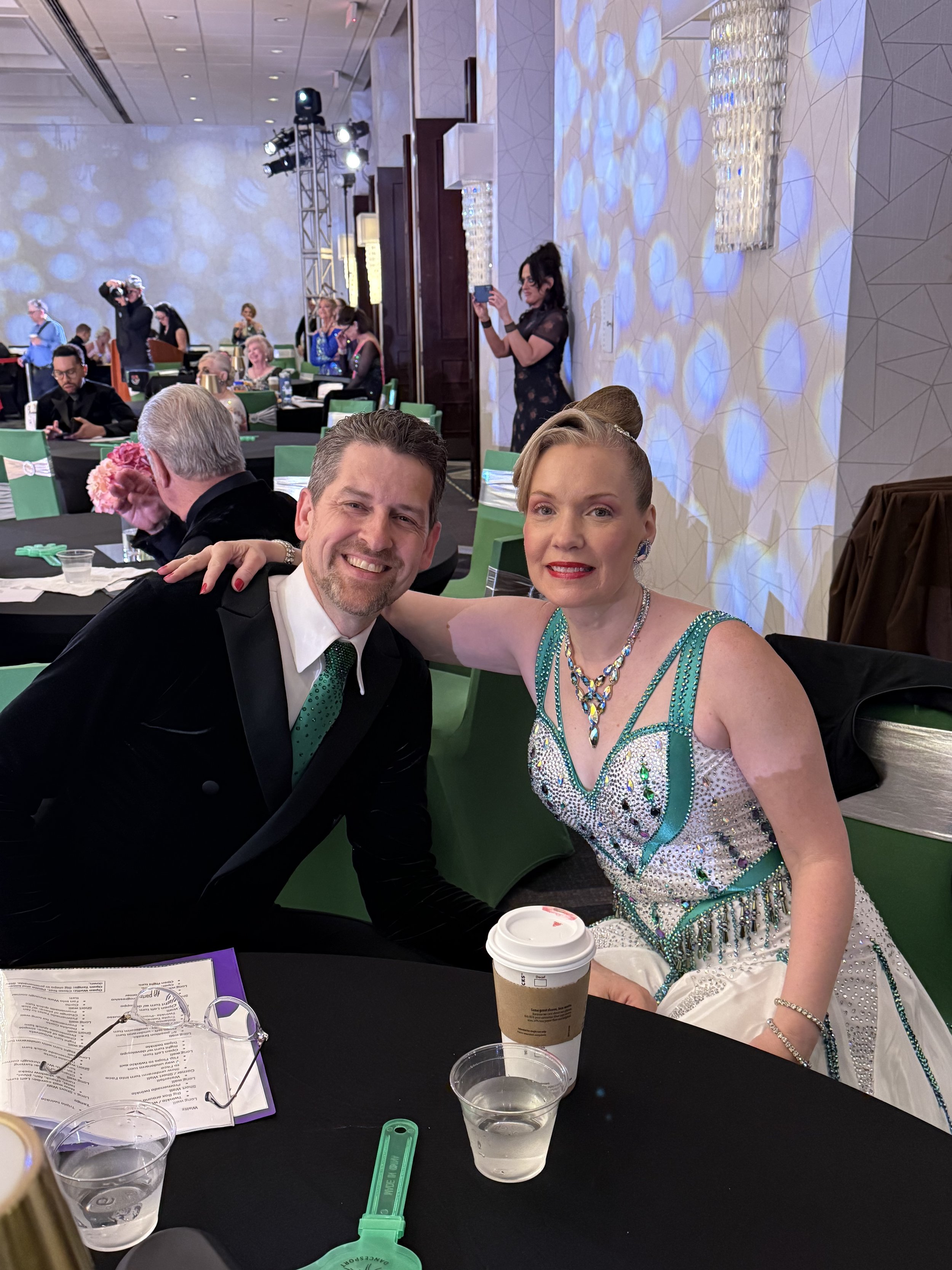 A man in a suit and a woman in a sparkling dress seated at a formal event, posing for a photo. They are at a table with drinks and papers. The background shows other people, event lighting, and decorations, suggesting a gala or ceremony atmosphere.
