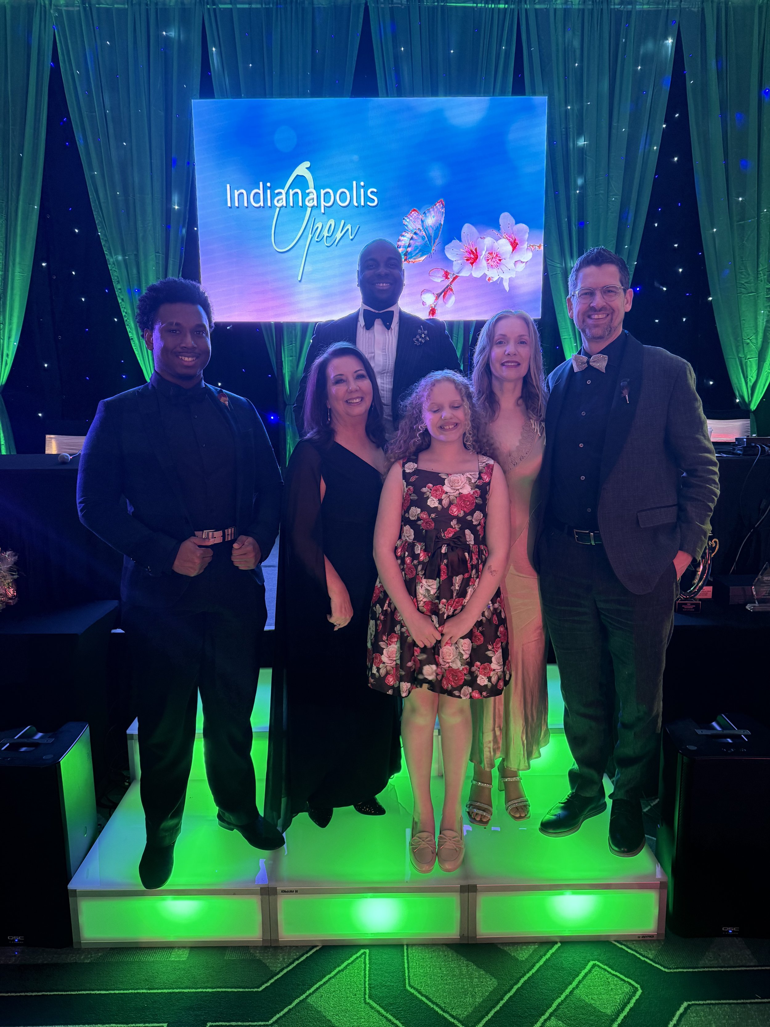 Group of six people in formal attire standing on a lit platform at the Indianapolis Open event with a blue and green backdrop.