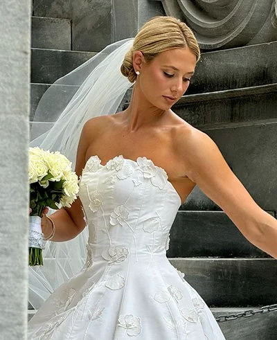 Bride in a white strapless wedding gown holding a bouquet of white roses, posing in front of a stone staircase.