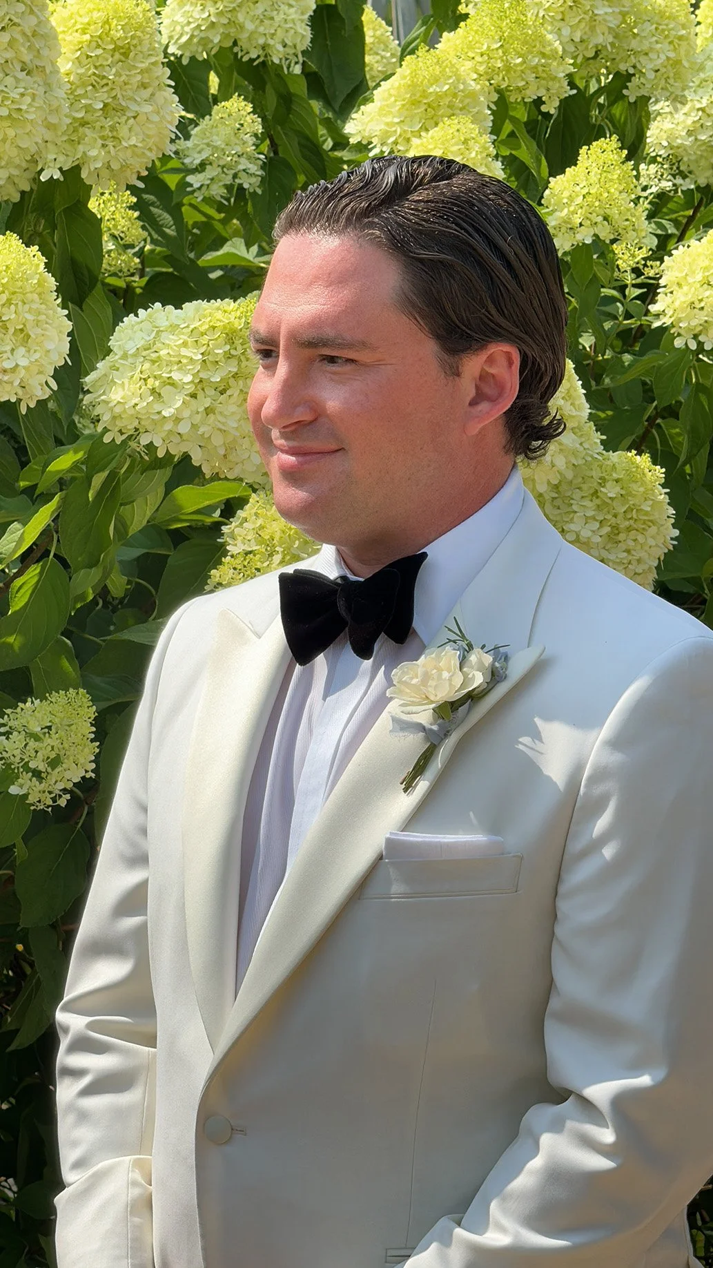 Man in a white tuxedo with black bow tie, standing in front of blooming hydrangea flowers.