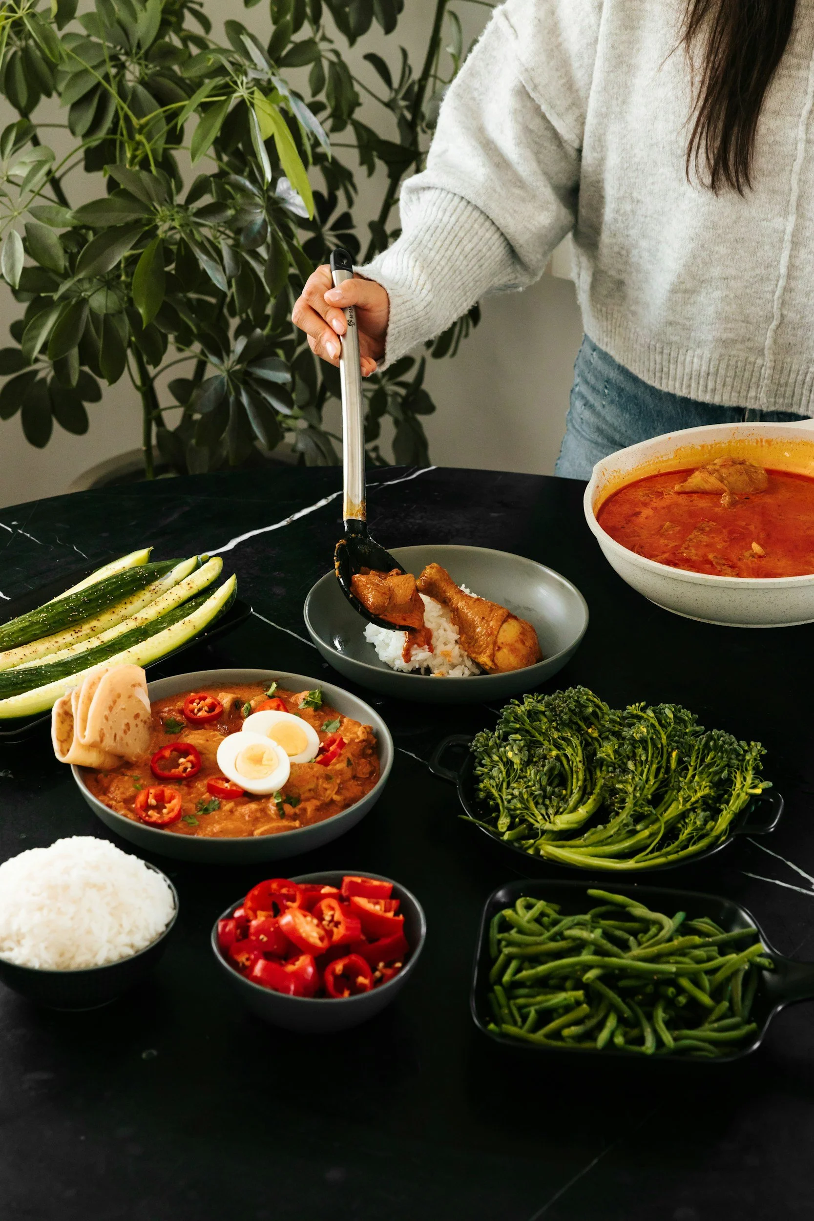 A table filled with various dishes including a bowl of white rice, a bowl of red chili or curry with chicken, a plate with chicken drumsticks over rice, noodles with sliced chili peppers and boiled egg, a bowl of sliced red chili, a plate of green beans, a bowl of broccoli, and cucumber slices. A person in a grey sweater is serving food onto their plate in a kitchen setting with a green leafy plant in the background.