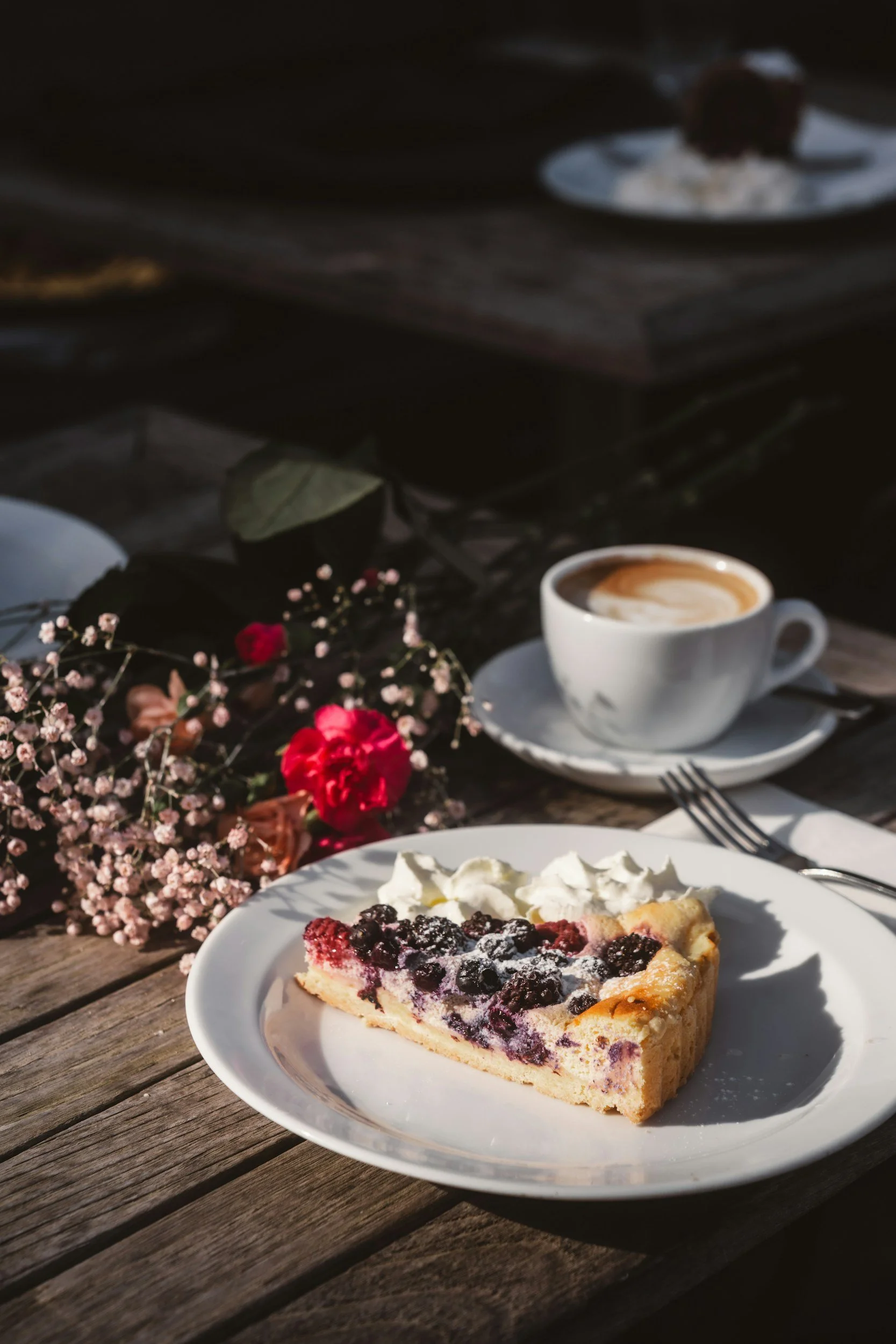 A slice of berry pie with whipped cream on a white plate, a cup of coffee, and flowers on a wooden table outdoors.