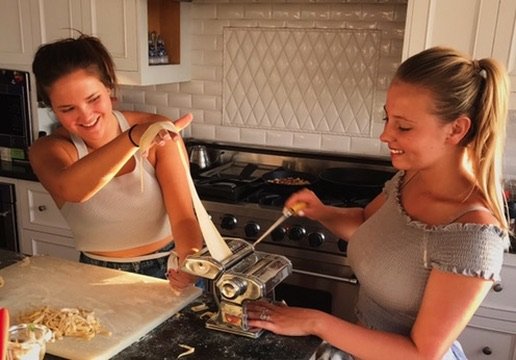 Two women in a kitchen making pasta together, with one pouring dough into a pasta machine and the other smiling as she assists.