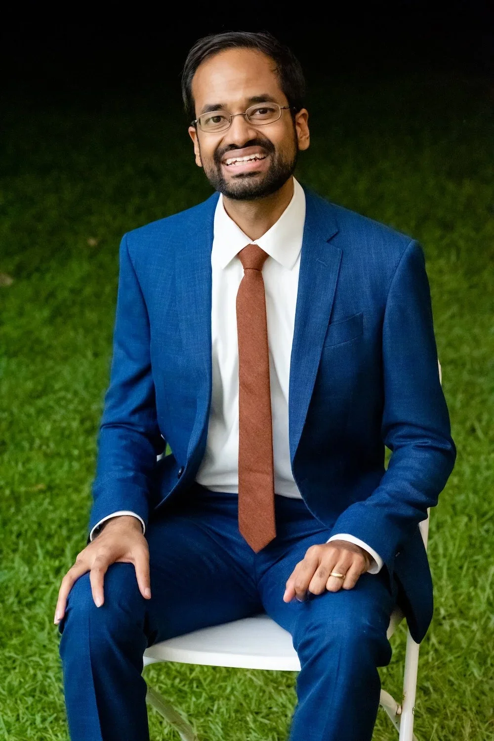 A man in a blue suit, white shirt, and brown tie sitting outdoors on a white chair on green grass, smiling at the camera.