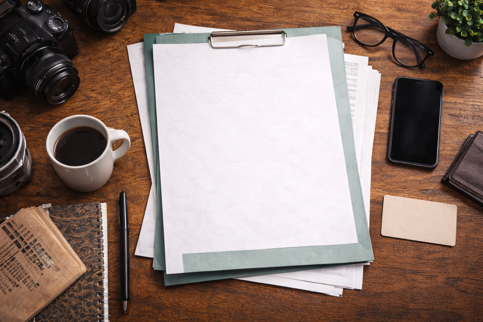 Organized desk with cameras, a cup of coffee, notebooks, papers, a pen, eyeglasses, a smartphone, a wallet, a plant, and a blank note card on a wooden surface.