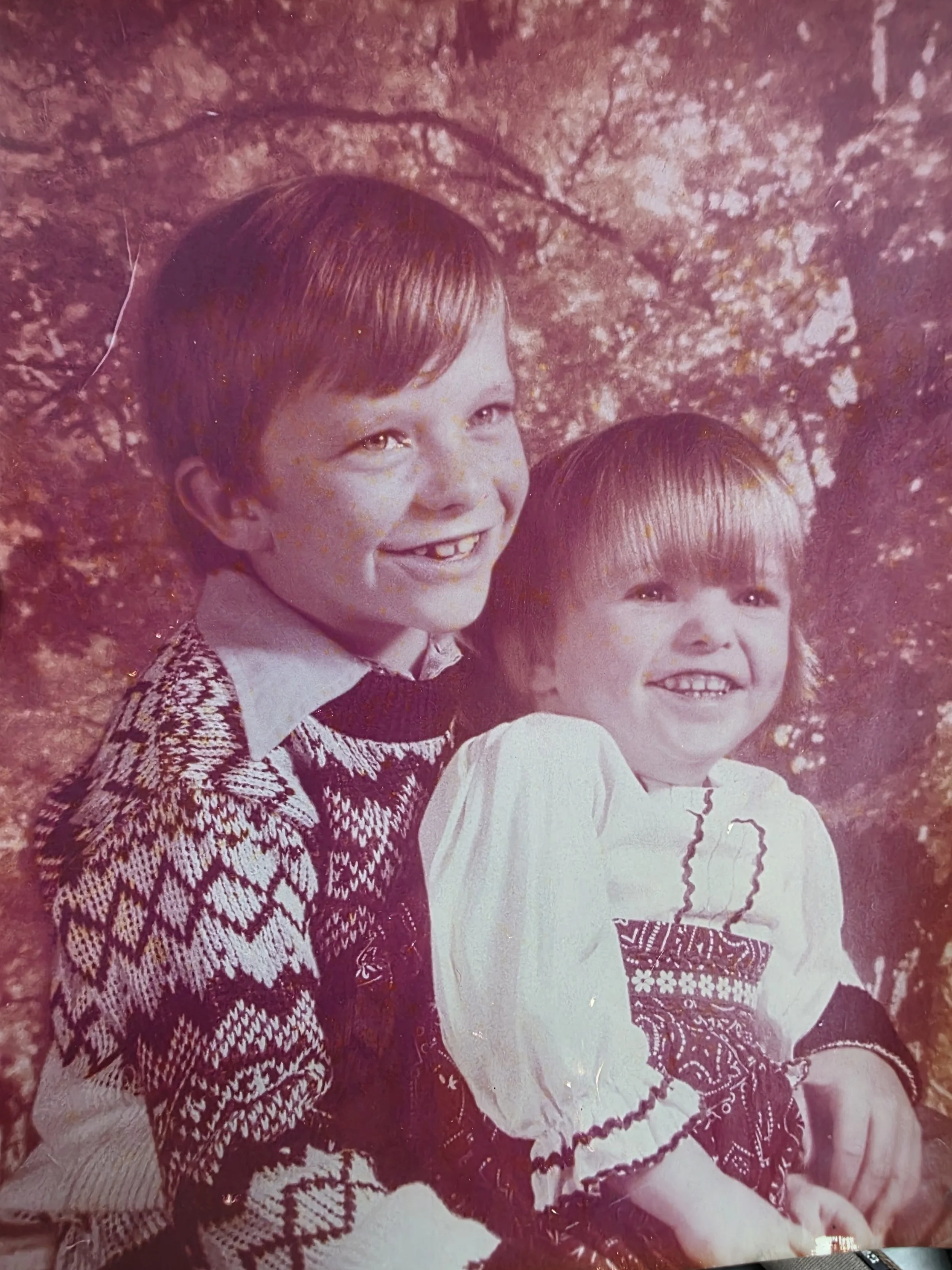 Two children, a boy and a girl, smiling and sitting outdoors with trees in the background. The boy has short red hair and is wearing a patterned sweater, while the girl has long straight hair and is wearing a knitted sweater with a heart design.