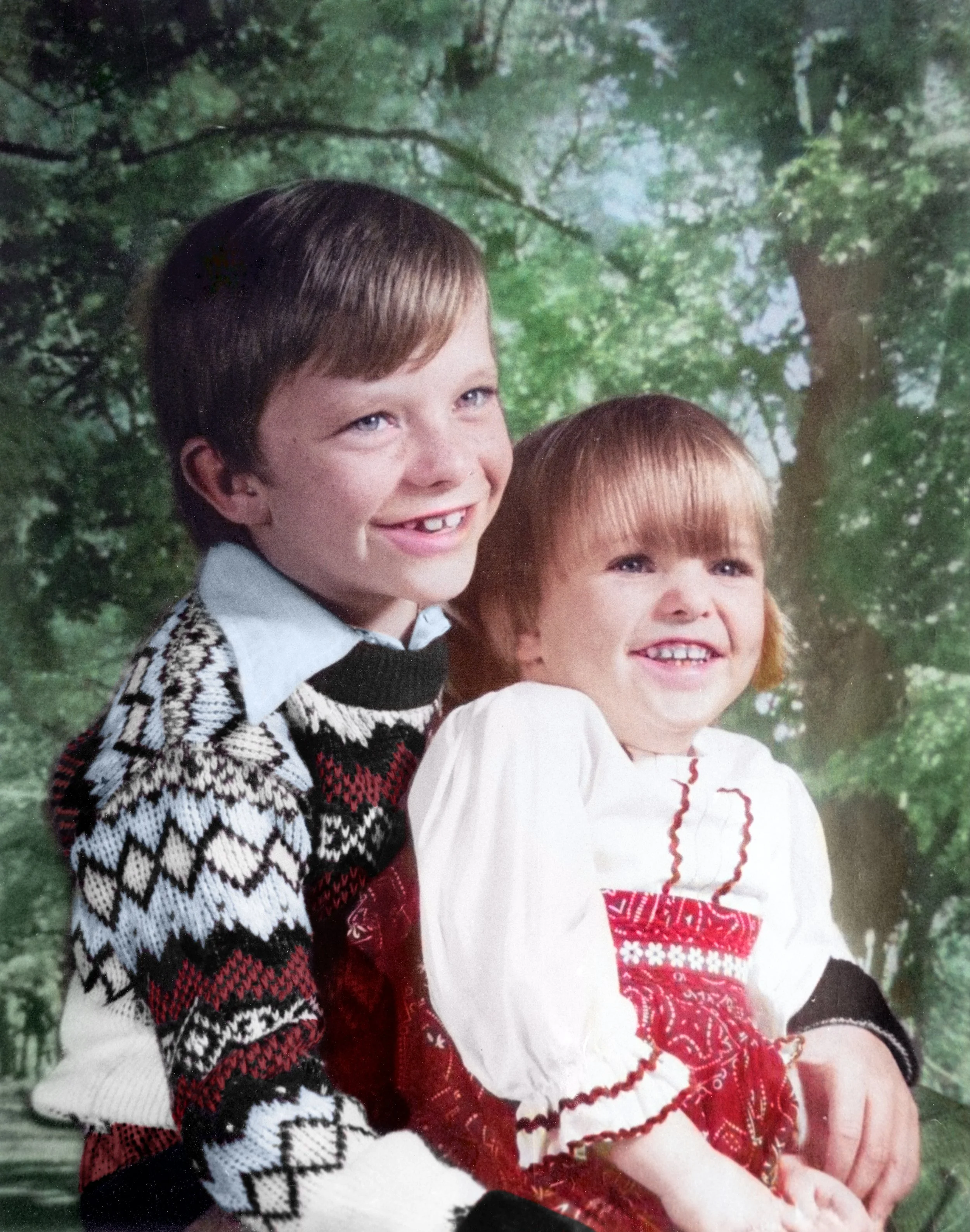 Two children, a boy and a girl, smiling outdoors with green trees in the background. The boy has short brown hair and wears a patterned sweater, the girl has reddish-brown hair and wears a Christmas-themed dress.