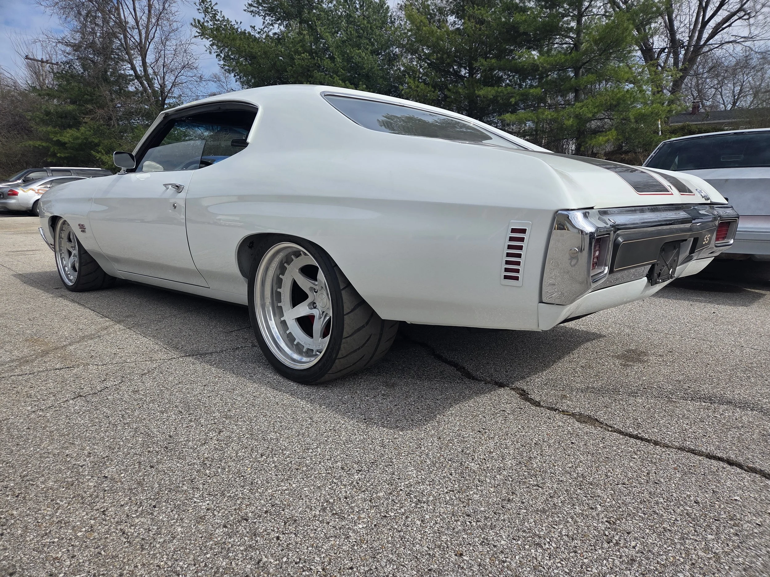 White vintage Chevrolet Chevelle SS muscle car with wide tires and custom rims parked on pavement, with trees and other cars in background.