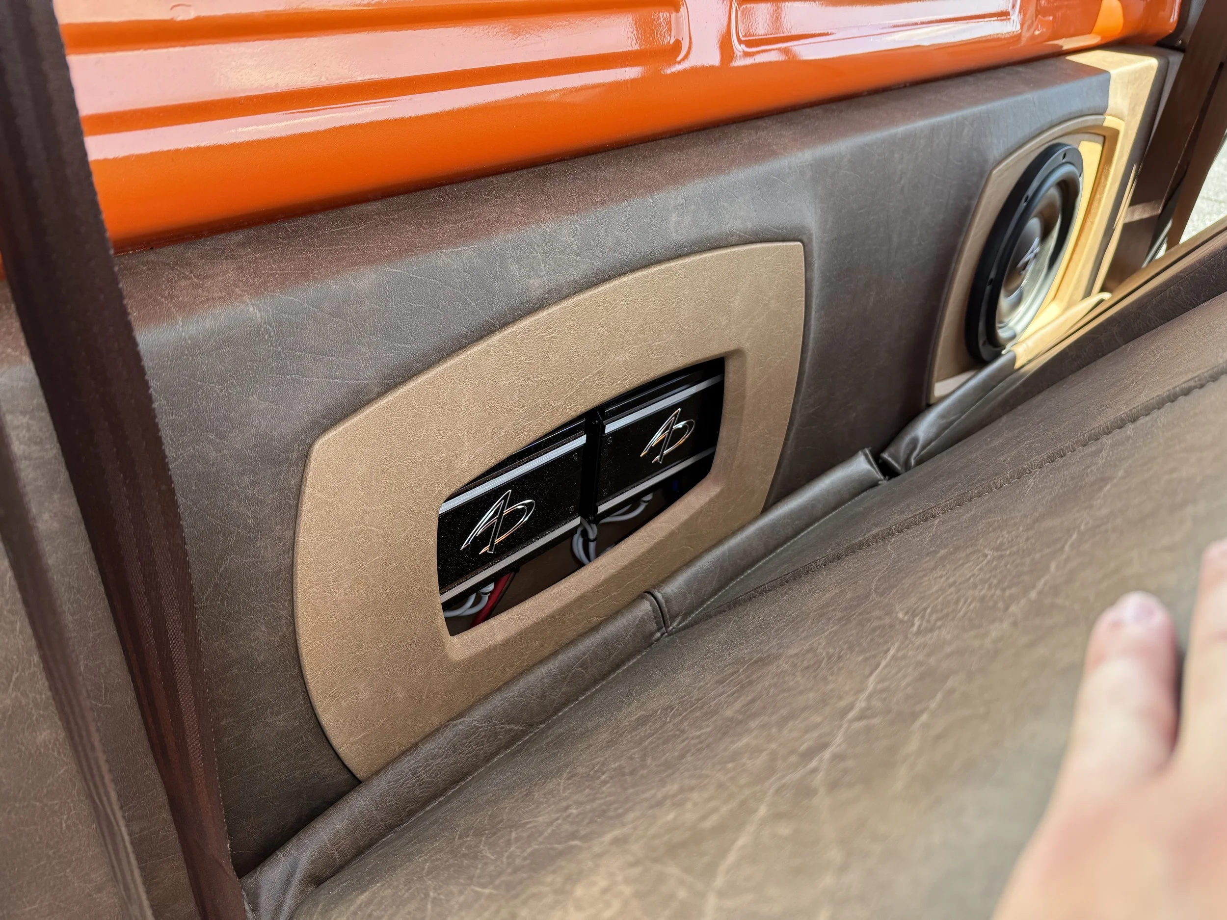 The back of a seat with brown upholstery and a pocket holding two Audio Dynamics-branded electronic devices, next to a speaker, with orange window trim above.