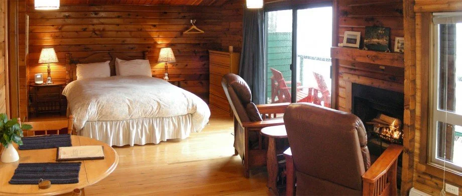 Cozy cabin bedroom with wooden walls, a large bed with white bedding, two bedside lamps, a sitting area with leather chairs, a fireplace, and a sliding glass door leading to an outdoor deck with chairs.