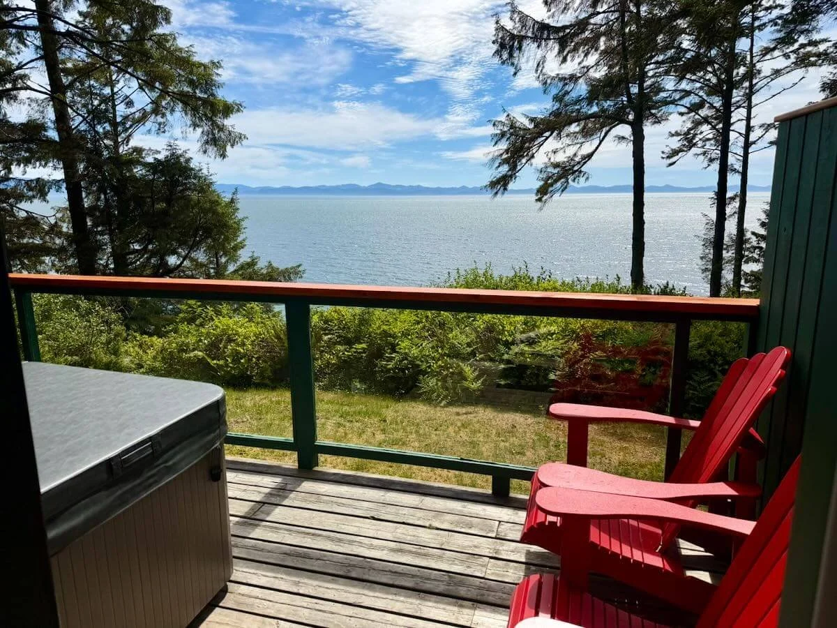 A view from a balcony overlooking a large body of water with distant mountains under a partly cloudy sky. The balcony has two red chairs, a small table, and a railing with greenery.