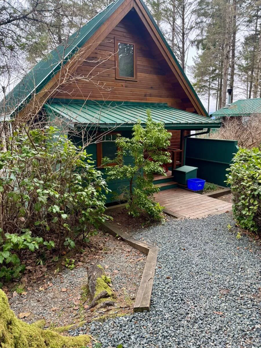 Cabin A exterior with a steep green metal roof, surrounded by trees and bushes, with a gravel pathway and a small wooden porch in a wooded area.