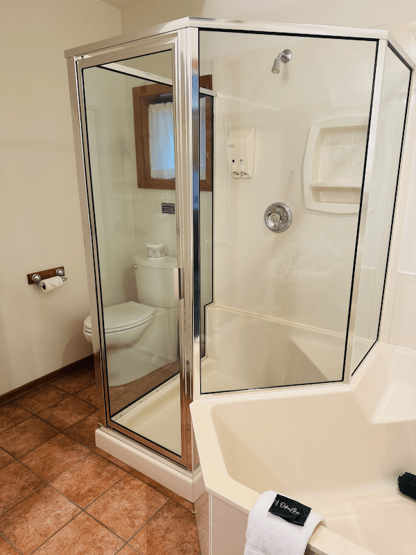 A bathroom with a corner shower enclosure with glass doors, a toilet, a small window with a curtain, and a bathtub with a towel on the edge.