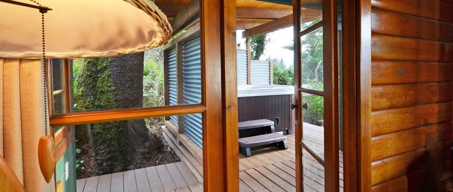 View from inside a wooden cabin looking out onto a deck with hot tub, trees, and outdoor landscape.