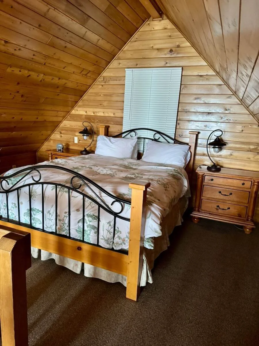 Cabin A's loft bedroom with wood-paneled sloped walls, featuring a queen-sized bed, two nightstands and lamps.