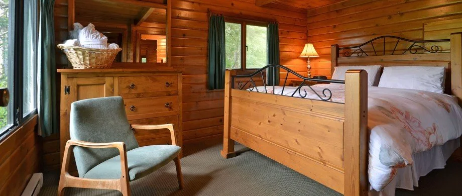A cozy bedroom with wooden paneled walls, a wooden bed with a metal headboard, a nightstand with a lamp, a chair with wooden armrests, and a dresser with a basket of towels near a window with green curtains.