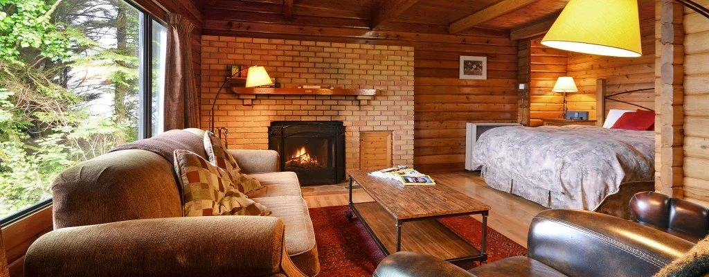 Cozy rustic living room with a large window showing trees outside, a brick fireplace, a sofa with pillows, a wooden coffee table, and a bed in the background.