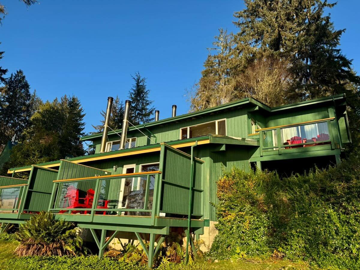 A green multi-level house on a hillside with a balcony and red outdoor chairs, surrounded by tall trees under a clear blue sky.