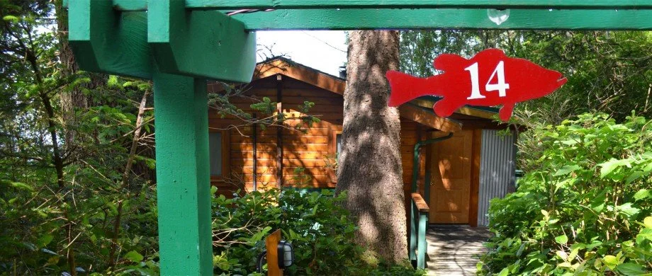 A red fish-shaped sign with the number 14 hangs from a green wooden structure. Behind it, there is a wooden cabin surrounded by trees and greenery.