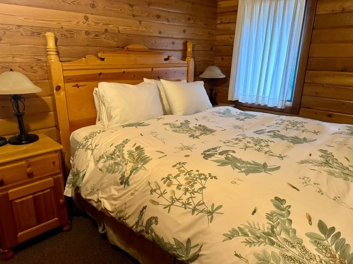 A queen-sized bed with white sheets and floral bedding, flanked by wooden nightstands with lamps, in a cozy wooden cabin bedroom.