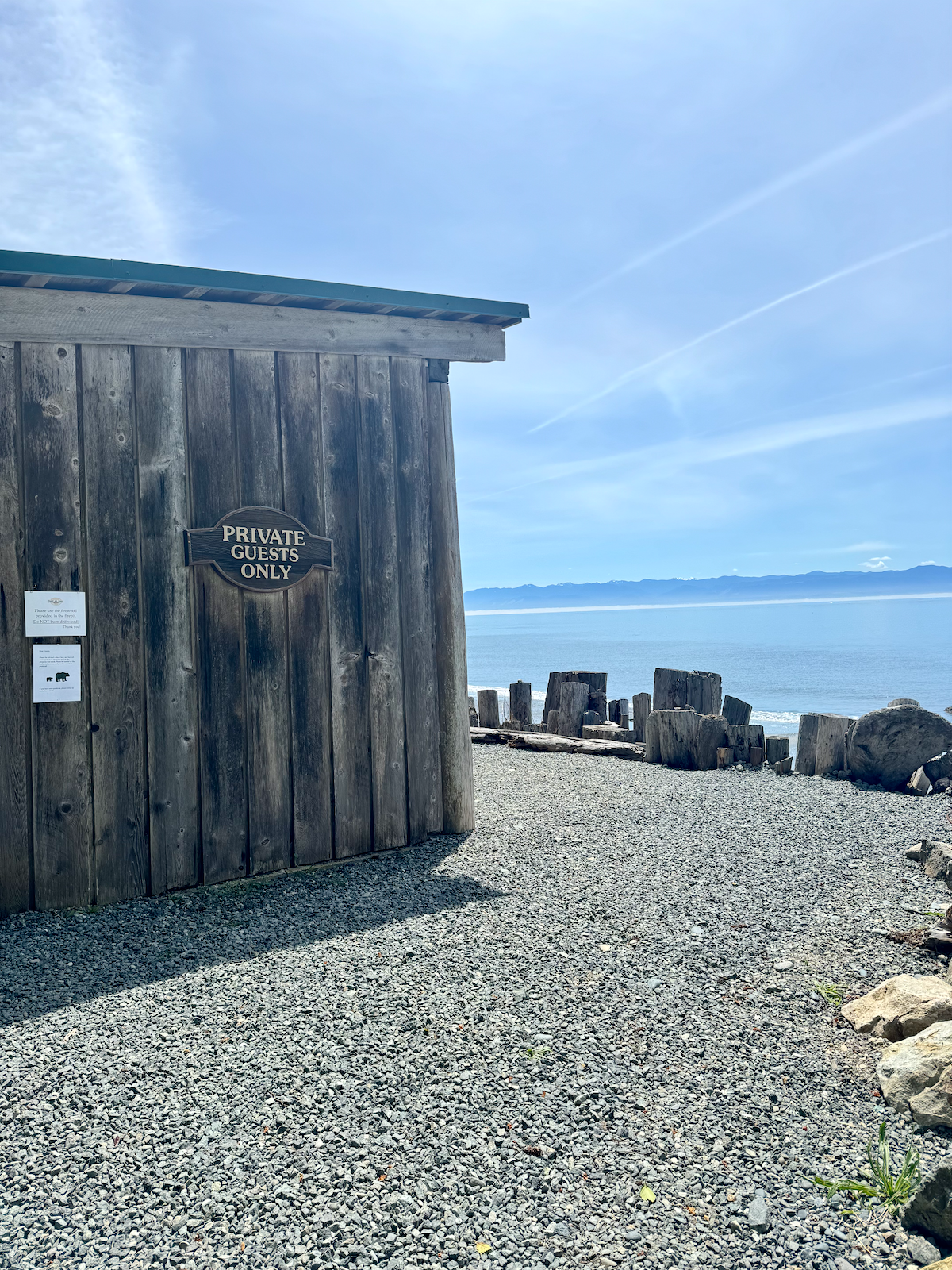 The Beach House with a sign that reads "Private Guests Only" near a rocky beach with driftwood logs and a view of the ocean and distant mountains under a blue sky.