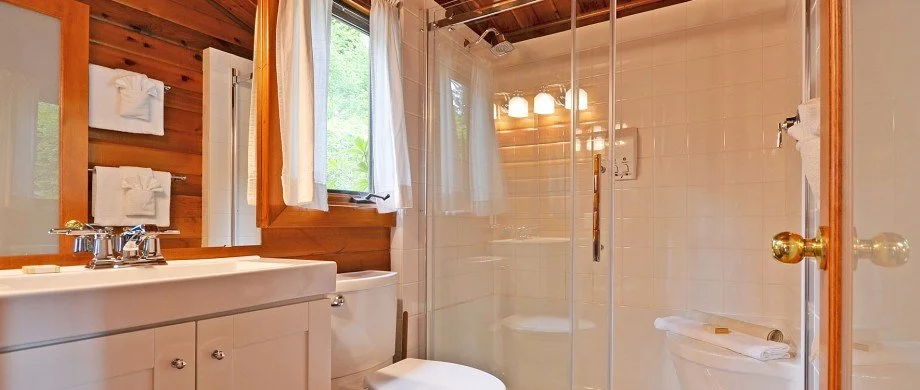Bathroom with wooden walls, white sink, mirror, window with curtains, toilet, and glass shower enclosure.