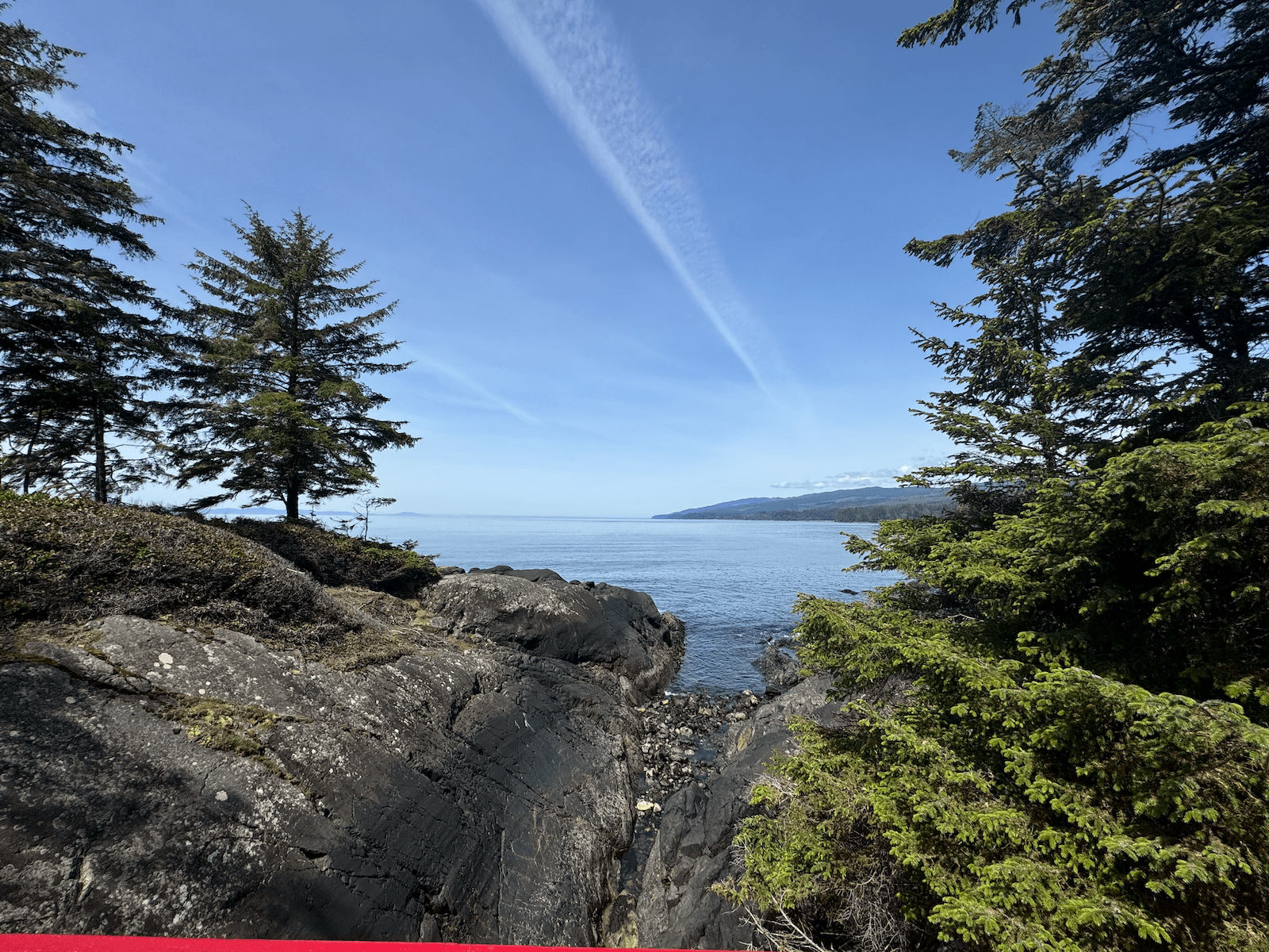 View of a rocky coastline with trees in the foreground, calm water, and a blue sky with wispy clouds.
