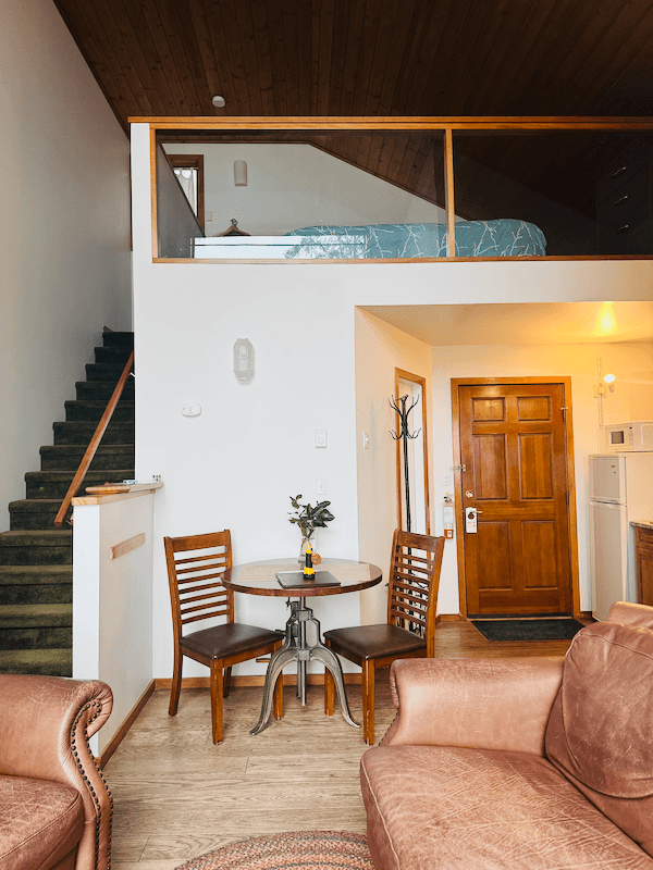 Cabin Blue Jay living room with a small round table and two wooden chairs, brown leather sofa in the foreground, staircase on the left, doorway with a wooden door in the background, and an upstairs loft with a visible bed and wooden railing.