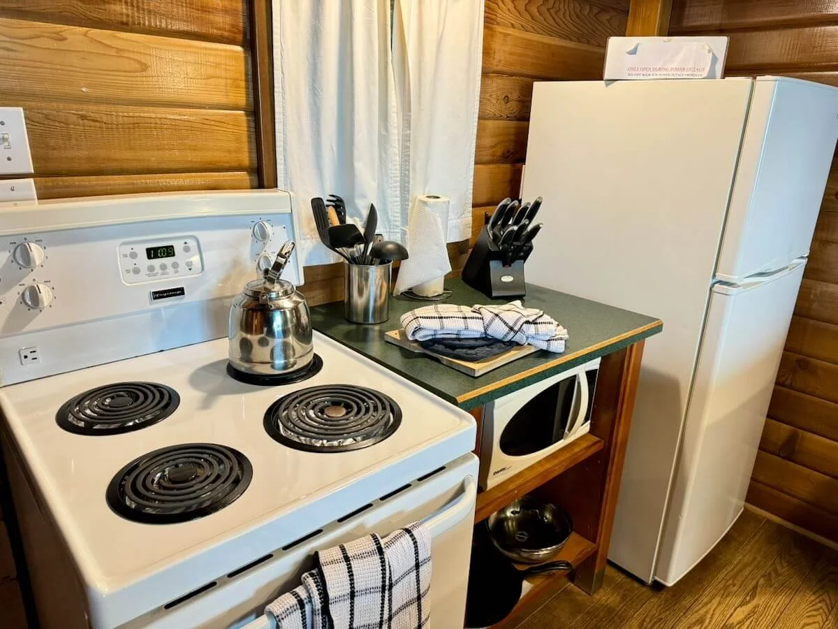 Cabin B kitchen with white electric stove, kettle, knives in a knife holder, paper towels, microwave, refrigerator, and wooden walls.