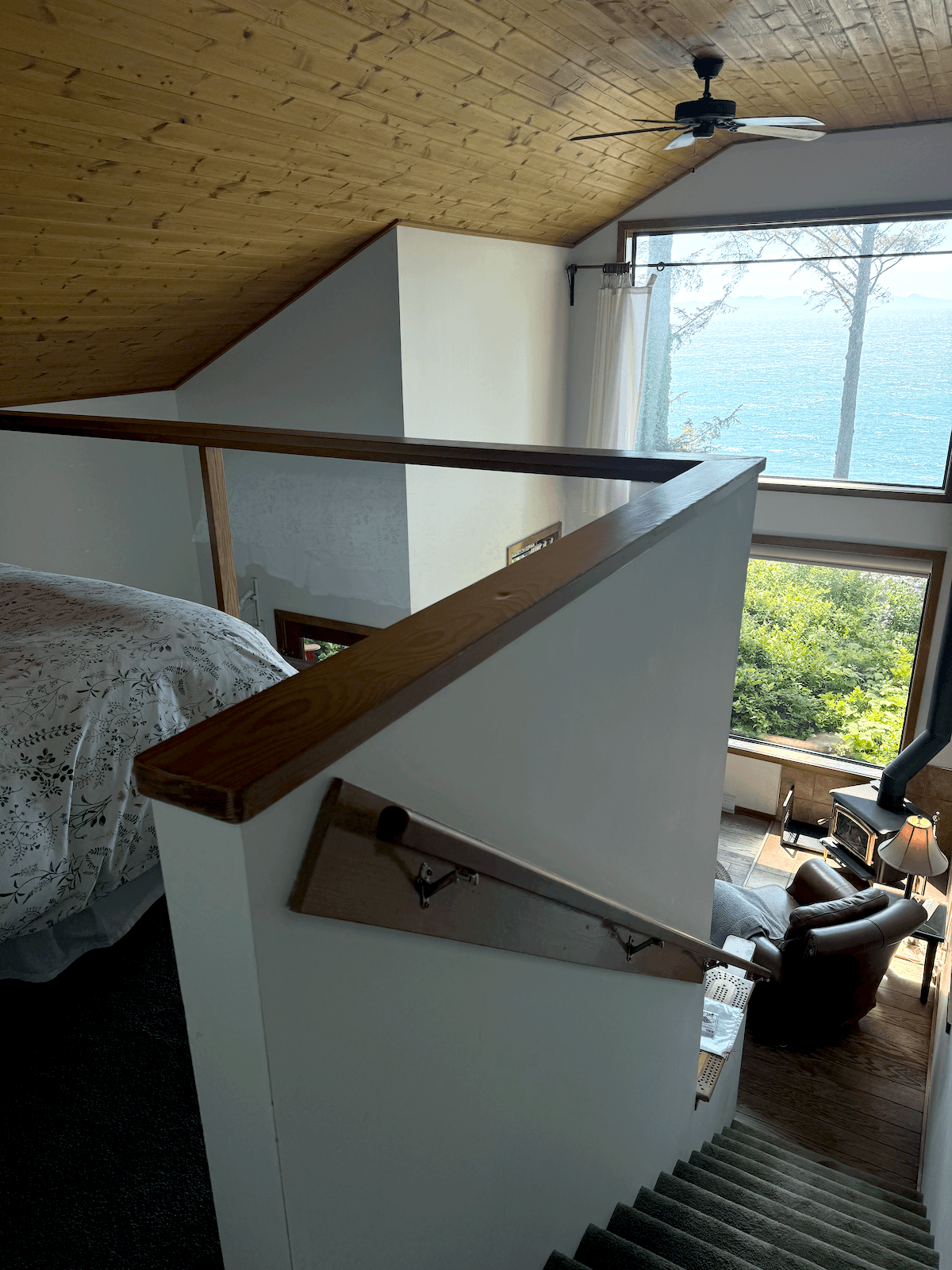 View of a the loft in Cabin Blue Jay, with large windows showing a lake and trees outside, a ceiling fan, and a cozy seating area with a fireplace.