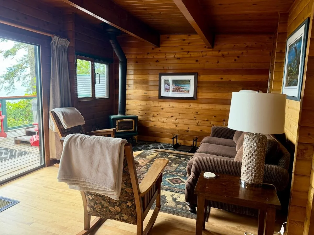 Cabin 10 living area with wooden walls, a brown sofa, a rocking chair with a blanket, a wood stove, and framed pictures on the wall. A sliding door opens to an outdoor deck with seating overlooking the ocean.