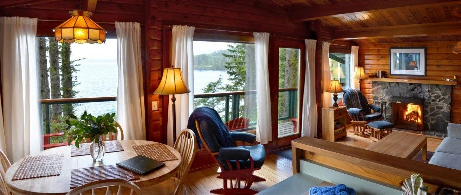 Interior of a cozy cabin living room with large windows showing a lake view, wooden walls, a stone fireplace, and various chairs and tables.