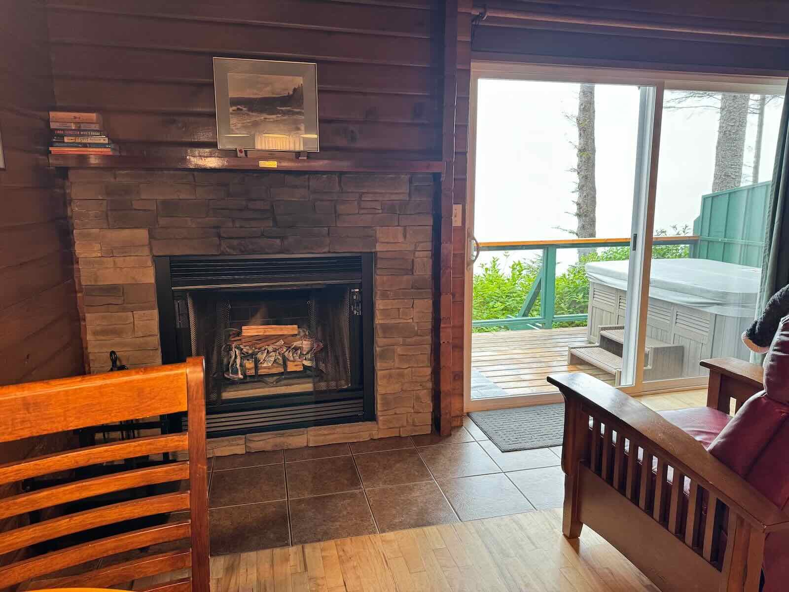 Cabin B interior with a wood-burning fireplace, wooden walls, and a sliding glass door leading to a deck with a hot tub. There are chairs, a small bookshelf, and a picture on the wall.