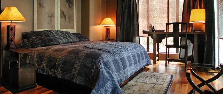 Bridge House bedroom with a king-sized bed, two bedside lamps, a wooden desk, a chair, and a rug, with curtains and sliding glass doors leading to an outdoor space.