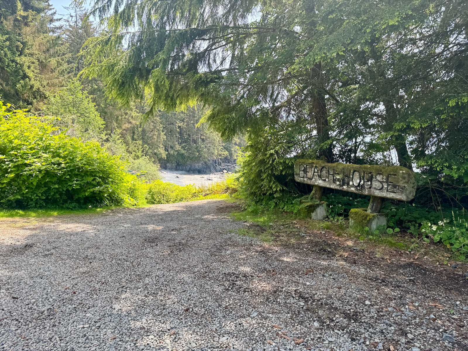 A gravel trail in a lush, green forested area leading to a rocky beach with water in the background. There is a weathered wooden sign that reads 'BEACH HOUSE' on the right side.