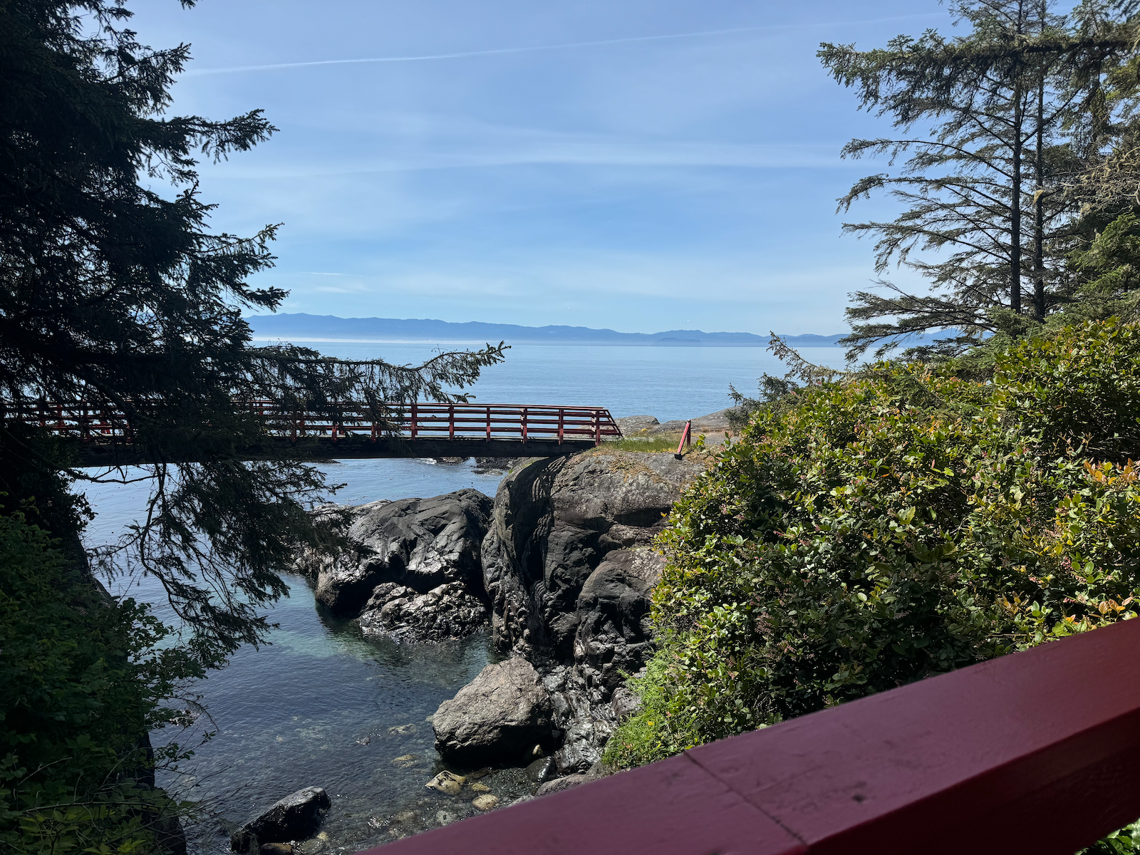 A scenic view of a body of water with a red bridge, rocky shoreline, green bushes, and tall trees on either side, with mountains and a clear blue sky in the background.