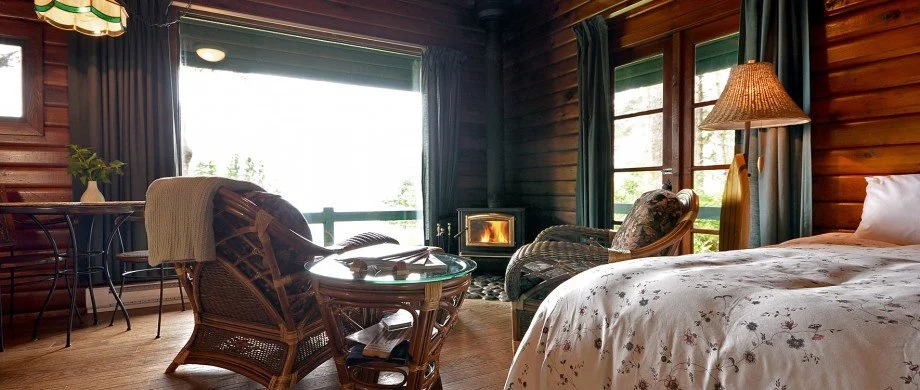 Cozy rustic bedroom with a wooden bed, floral bedding, a small fireplace, wicker chairs, a side lamp, and large windows with curtains in a log cabin.
