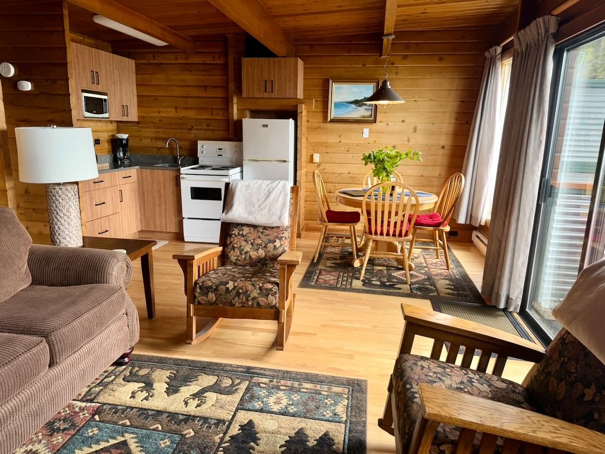 Cabin 10 interior view with a small kitchen, sitting area, and dining table. The kitchen has a microwave, coffee maker, stove, and refrigerator. The sitting area features a sofa, armchairs, a side table, and a lamp. 