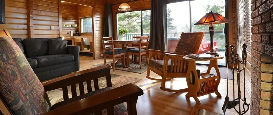 Bridge House living room with wooden walls and large windows, furnished with a black sofa, a wooden rocking chair, a small table with a lamp, a dining table with four chairs, and a brick fireplace.