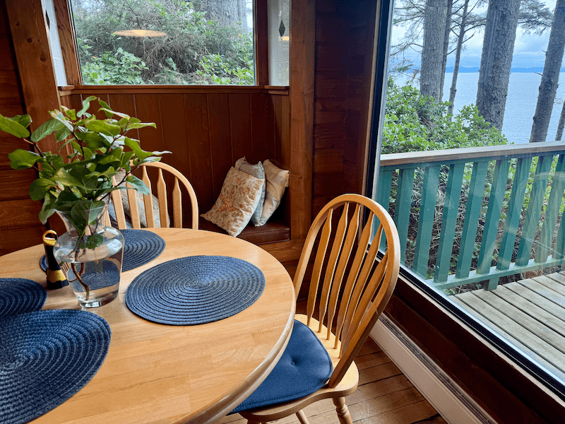 A cozy corner of a cabin with a wooden round dining table and three blue placemats. A small glass vase with green leaves sits on the table. Two wooden chairs with blue cushions are around the table. A built-in bench by the window has pillows. Large glass window and door reveal a balcony with a wooden railing and a view of trees and water outside.