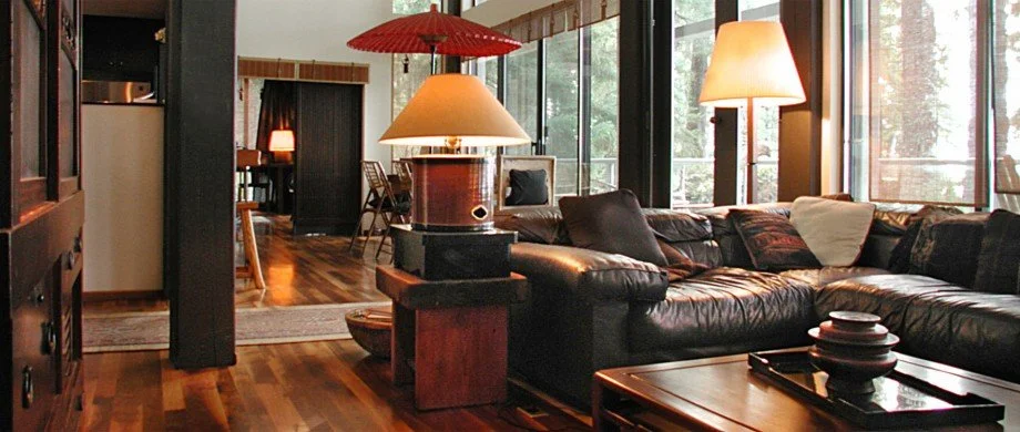 A cozy living room with a large black leather sectional sofa, wooden coffee table, floor lamps, and large windows providing natural light.