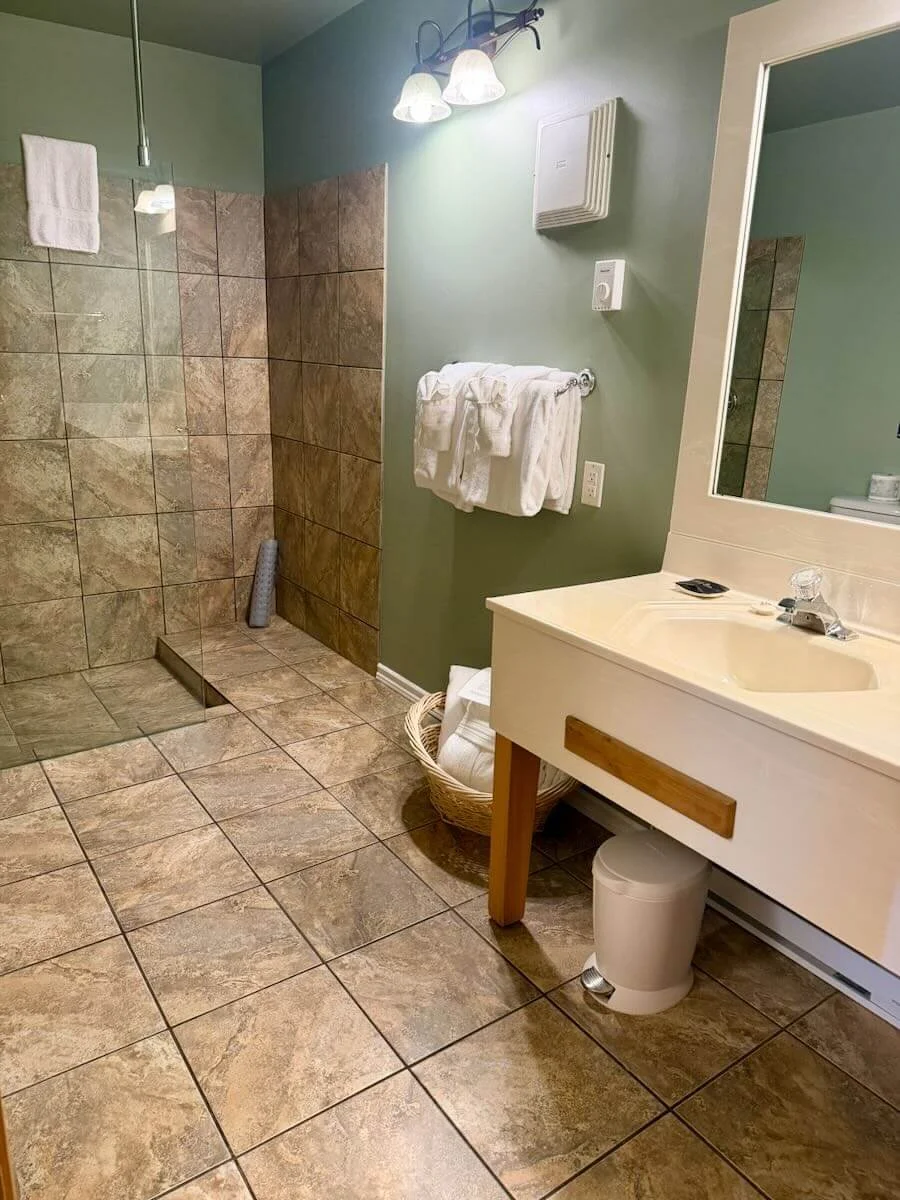 A bathroom with a tiled walk-in shower, towel rack with white towels, a basket with towels, a sink with a mirror above, and a trash bin underneath